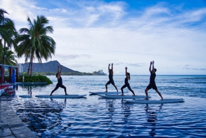 ダイヤモンドヘッドを望む海辺で、穏やかな朝日を浴びながらボードの上でポーズをとる人々。