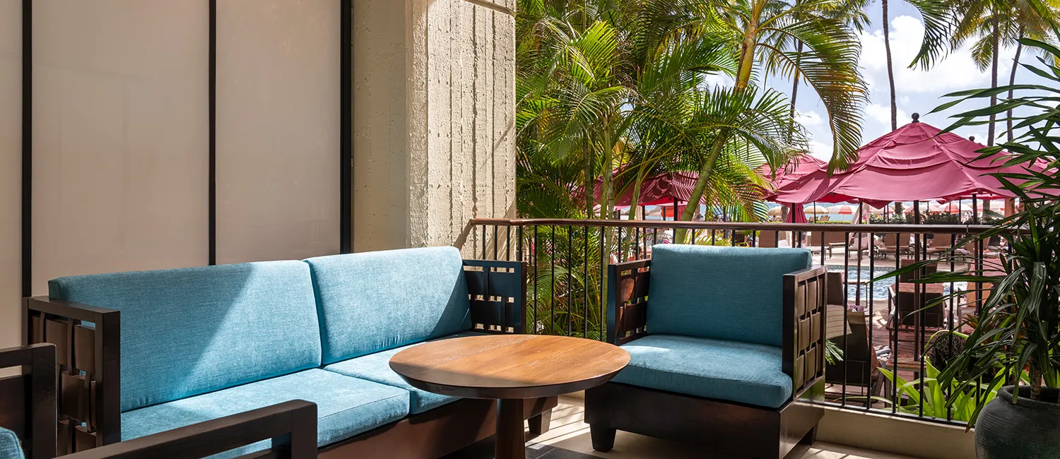 バルコニーにブルーのソファと木製テーブルを備え、ピンクのパラソルが並ぶプールエリアを望む