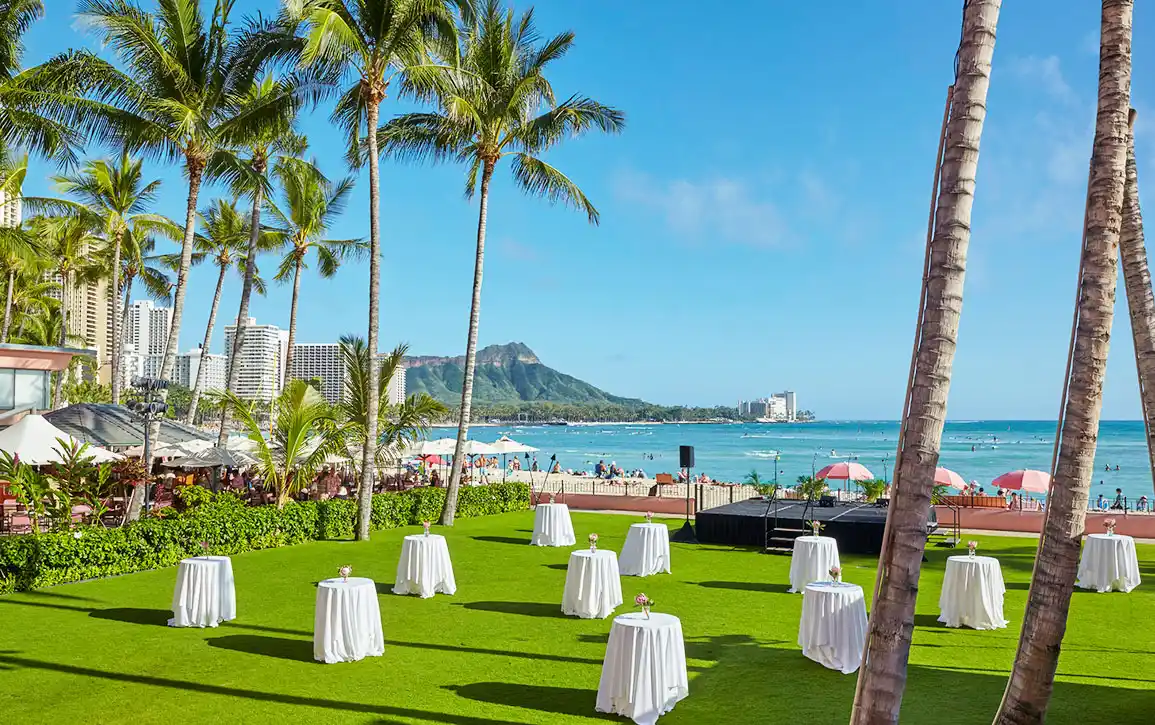 ヤシの木に囲まれた芝生に、白いクロスのハイテーブルが配置されたオーシャンフロントのレセプションスペース
